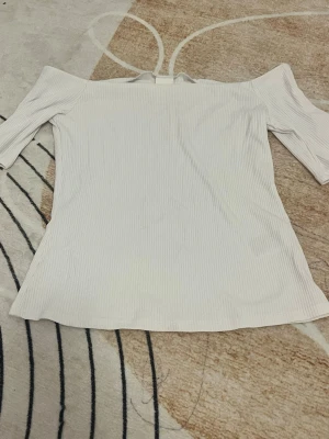 Vit ribbad offshoulder topp - Säljer en vit ribbad topp med offshoulder-design och trekvartsärm. Toppen har en stilren och enkel look med smal ribbad struktur i tyget, perfekt för att skapa en fräsch och modern outfit.