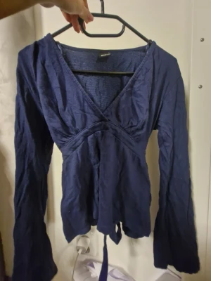 Mörkblå omlottopp med knyt - Säljer en mörkblå omlottopp från Ginatricot med långa ärmar och v-ringad hals. Toppen har ett band i midjan som knyts baktill för en snygg siluett. Tillverkad i ett mjukt och lätt bomullstyg som känns skönt mot huden. Skrynklig pg av att den låg i garderoben länge utan användning.