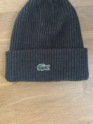 Mörkgrå ribbad mössa från Lacoste - Säljer en mörkgrå ribbstickad mössa från Lacoste med klassisk krokodillogga framtill. Kom med prisförslag, skriv för fler bilder :)