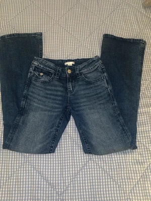 Blå jeans med rosett från Gina Tricot - Snygga blå jeans från Gina Tricot i klassisk femficksmodell. Baktill finns två fickor med broderade kors och dekorativa knappar. Jeansen har normal passform och är tillverkade i slitstarkt denimtyg. Perfekt för dig som gillar detaljer och vill sticka ut.