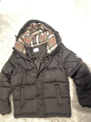 Burberry puffer jacket  - Svart pufferjacka från Burberry med stor huva och tydlig logga på ärmen. Insidan har ett rutigt foder i grått, vitt och rött. Perfekt för kalla dagar och riktigt snygg streetstil. Aldrig använt!!!!! 