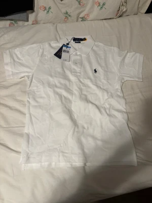 Vit pikétröja från Ralph Lauren - Klassisk vit pikétröja från Polo Ralph Lauren i bomull med korta ärmar och krage. Tröjan har två knappar framtill och den ikoniska mörkblå broderade loggan på bröstet. Classic fit-modell som passar till allt.