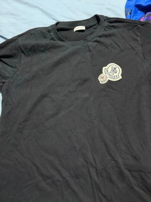 Svart Moncler t-shirt med logga - Svart t-shirt från Moncler med rund hals och korta ärmar. På bröstet sitter en snygg broderad Moncler-logga i vitt och blått. T-shirten är gjord i mjuk bomull och har en klassisk passform som funkar till många olika stilar.