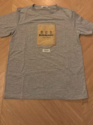 Grå Burberry t-shirt med beige patch - Säljer en grå t-shirt från Burberry i mjuk bomull. Framtill finns en stor beige patch med Burberry-logga och tryck i svart, samt en mindre Burberry-label under. Klassisk rund hals och korta ärmar. Perfekt för dig som gillar stilrena designer med en twist.