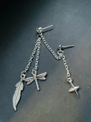 By prisförslag- 1 st Silvrig kedjeörhänge med berlocker - Unikt örhänge i silverfärgad metall med kedjor och tre berlocker: en fjäder, en trollslända och en stjärna. Snygg detalj till din stil och passar perfekt för dig som gillar accessoarer med lite edge.