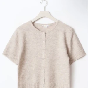 Beige stickad kofta från Gina Tricot - Kortärmad, beige stickad kofta från Gina Tricot med rund halsringning och knappar framtill. Mjuk och fluffig känsla som är stilren och passar till nästintill allt! Passar perfekt för dig som vill vara bekväm i vardagen men också vara lite mer finklädd. 