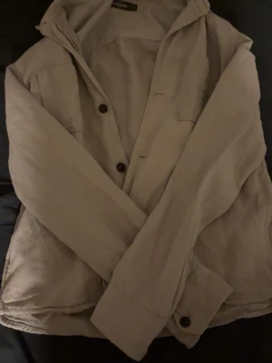Beige overshirt från Dressmann - Snygg beige overshirt från Dressmann i storlek S. Jackan har en klassisk krage, bröstficka och knäppning med svarta knappar. Tillverkad i mjukt bomullsmaterial, perfekt att slänga på över en t-shirt eller tröja. Enkel och stilren design som funkar till jeans.