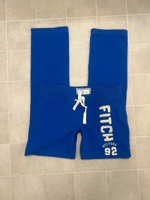 Blå mjukisbyxor från Abercrombie & Fitch - Snygga blå lågmidjade mjukisbyxor från Abercrombie & Fitch med vit snörning i midjan och broderad text 'FITCH 92 NEW YORK' på benet. Storlek M.
