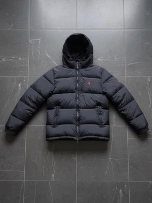 Svart pufferjacka från Polo Ralph Lauren - Svart pufferjacka från Polo Ralph Lauren med huva och dragkedja framtill. Jackan har en röd logga på bröstet och quiltad design. Materialet är polyester och dun, perfekt för kalla dagar. Snygg och stilren modell som håller värmen.