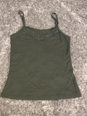 Olivgrön linnetopp med spets - Säljer en olivgrön linnetopp med smala axelband och dekorativ spets längs halsringningen. Toppen är ärmlös och har en enkel, rak passform. Perfekt för varma dagar och enkel att matcha med jeans eller shorts.