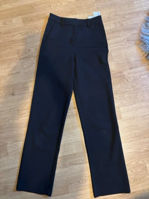 Svarta kostymbyxor med raka ben - Säljer ett par svarta kostymbyxor med klassisk rak passform och hällor för bälte. Byxorna har diskreta fickor och är perfekta för en clean och stilren look. Materialet känns mjukt och följsamt, vilket gör dem riktigt sköna att bära.