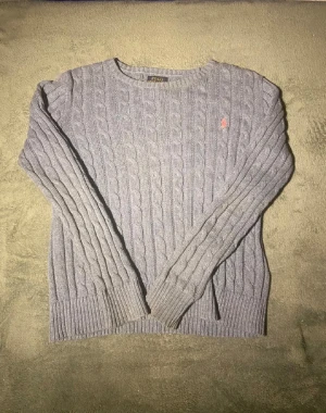 Grå kabelstickad tröja Polo Ralph Lauren - Snygg grå kabelstickad tröja från Polo Ralph Lauren i storlek L. Tröjan har lång ärm, rund hals och klassisk röd broderad logga på bröstet. Ribbstickade muddar vid ärmslut och nederkant ger en schysst passform. Perfekt för dig som gillar stilrena och tidlösa plagg.