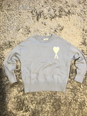 Ljusblå stickad tröja från Ami - Säljer en ljusblå stickad tröja från Ami Alexandre Mattiussi med ett vitt hjärta och A broderat på bröstet. Tröjan har rund hals, ribbade muddar och är långärmad. Perfekt för dig som gillar stilrena detaljer och mjuka material.