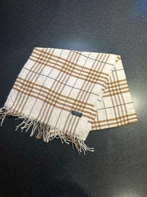 Rutig ullväst från Burberrys - Snygg väst från Burberrys i klassiskt rutigt mönster med beige och bruna toner. Västen är tillverkad i 100% lammull och har fransar längs nederkanten. Perfekt för dig som gillar tidlös design och vill ha något som sticker ut.