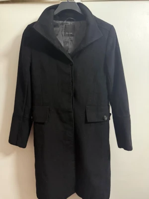 Svart kappa från Sand i kashmirull - Svart kappa från Sand med klassisk krage och knappar framtill. Tillverkad i en lyxig blandning av kashmir och ull. Snygga fickor med lock och knappdetaljer på ärmarna. Perfekt för dig som vill ha en stilren och tidlös look.