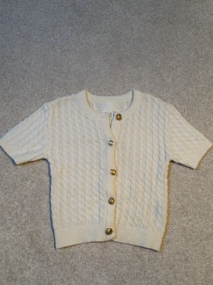 Kortärmad kabelstickad kofta från H&M, nyskick. - Snygg kabelstickad kofta från H&M i offwhite med korta ärmar och rund hals. Koftan har fem guldfärgade knappar framtill och ribbade muddar vid ärmslut och nederkant. Perfekt för gillar en elegant och lyxig stil.