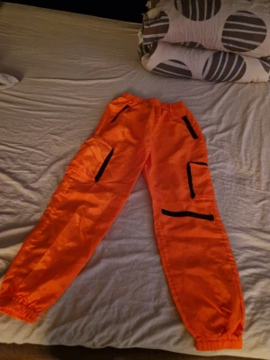 Orange cargopants med svarta detaljer - Snygga orange cargopants med flera svarta dragkedjor och fickor på benen. Byxorna har resår i midjan och vid bensluten för en skön och avslappnad passform. Perfekta för dig som vill sticka ut med färg och funktionella detaljer.