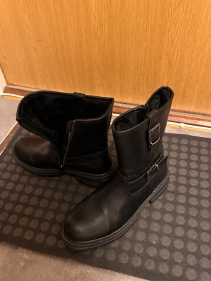 Svarta skinnstövlar med spännen - Snygga svarta stövlar i skinn med två dekorativa spännen och dragkedja på sidan. Insidan är fodrad för extra värme och komfort. Perfekta för kalla dagar och ger en cool look till din outfit.