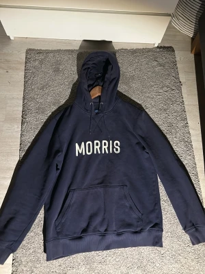 Marinblå hoodie från Morris - Snygg marinblå hoodie från Morris med stor vit logga på bröstet. Klassisk känguruficka framtill, ribbade muddar och justerbar huva med dragsko. Perfekt för en chill och avslappnad stil. Mjuk bomullskänsla och riktigt skön passform. Storlek M. Bara att höra av er om frågor och funderingar🙌🙌