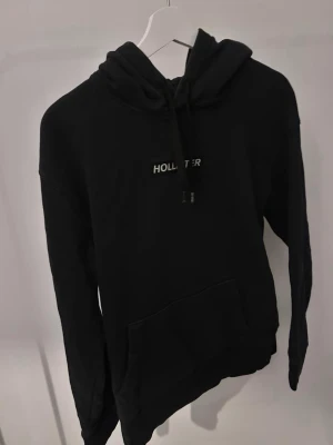 Svart hoodie från Hollister - Snygg svart hoodie från Hollister med klassisk logga på bröstet. Hoodien har huva med dragsko och en stor magficka framtill. Tillverkad i mjukt bomullsmaterial som är skönt att ha på sig. Perfekt för en avslappnad och cool stil.
