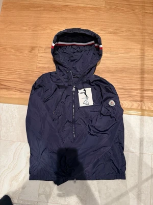 Moncler windbreaker  - Snygg marinblå vindjacka från Moncler med huva och dragkedja framtill. Jackan har Moncler-logga på ärmen och rödvit detalj på huvan. Tillverkad i lätt och glansig polyester, perfekt för blåsiga dagar.