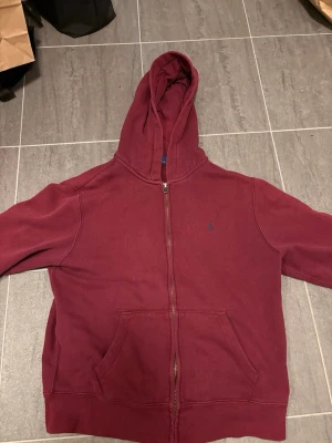 Vinröd hoodie från Polo Ralph Lauren - Snygg vinröd hoodie med dragkedja från Polo Ralph Lauren. Klassisk design med huva, kängurufickor och broderad blå logga.