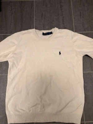 Vit tröja från Polo Ralph Lauren - Klassisk vit tröja från Polo Ralph Lauren med rund halsringning och den ikoniska broderade loggan på bröstet. Tröjan har ribbade muddar vid ärmslut och nederkant. Perfekt för en stilren look.