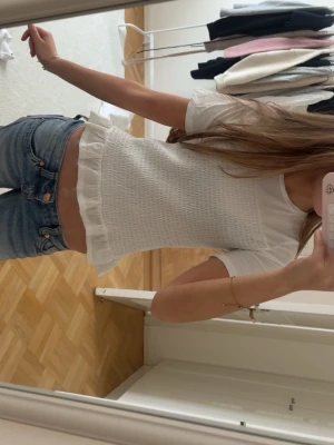 Vit blus med volangkant - Söt vit blus med smockad front och volangkant nertill. Toppen har korta ärmar och rund halsringning, vilket ger en feminin och fräsch look. Perfekt att matcha med jeans eller kjol för en trendig stil.