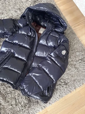 Mörkblå dunjacka från Moncler - Snygg mörkblå pufferjacka från Moncler med glansig finish och huva. Jackan har dragkedja framtill, fickor med dragkedja och klassisk Moncler-logga på ärmen. Perfekt för kalla dagar när du vill hålla stilen.