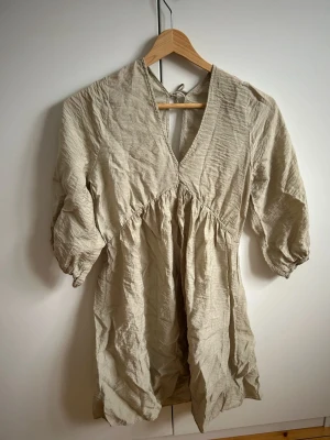 Khaki strand/sommarklänning med v-ringning och knyt - Luftig Khaki strand/sommarklänning med v-ringning och knyt i nacken. Blusen har trekvartsärm. Skrynklig på bilden men försvinner efter en tvätt! Skickar snabbt