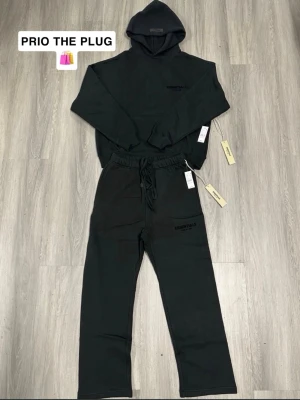 Svart Essentials tracksuit  - Svart loungewear set från Essentials med hoodie och matchande byxor. Setet har minimalistisk design, diskret logga på både tröja och byxa samt mjukt bomullsmaterial. Hoodien har stor huva och byxorna har snörning i midjan för extra komfort.