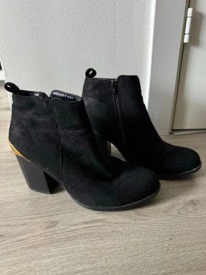 Svarta stövletter med klack - Snygga svarta stövletter i mocka-look med bred klack och gulddetalj vid hälen. Dragkedja på insidan för enkel på- och avtagning. Perfekta för att lyfta vilken outfit som helst.