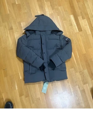 Canada Goose wyndham grå black label - Snygg mörkgrå dunjacka från Canada Goose med stor huva och patch på ärmen. Jackan har långa ärmar, dragkedja och knappar framtill samt en quiltad design som håller värmen under kalla dagar. Perfekt för vintern och riktigt stilren.