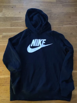 Svart Nike hoodie med stor logga - Svart hoodie från Nike med stor vit logga och swoosh på bröstet. Klassisk känguruficka framtill och justerbar huva med dragsko. Tillverkad i mjukt bomullsmaterial som är skönt mot huden. Perfekt för en avslappnad och sportig stil.