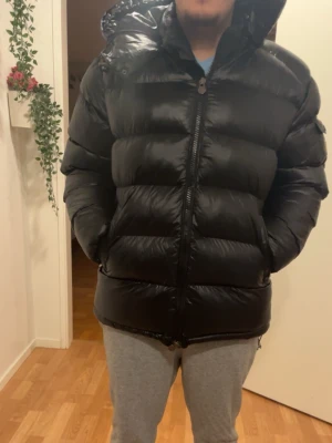 Svart Moncler pufferjacka  - Säljer en svart pufferjacka från Moncler med klassisk logga på ärmen. Jackan har quiltad design, dragkedja framtill och justerbar snörning nedtill. Materialet är glansig polyester och dunfyllning, perfekt för kalla dagar. Insidan har en unik Moncler-serieetikett. Den går att skanna 