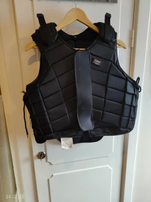 Svart skyddsväst från CRW - Säljer en svart skyddsväst från CRW med vadderad design och justerbara axelband. Västen har rutmönstrad struktur och bred kardborrestängning framtill. Perfekt för ridning eller annan sport där extra skydd behövs.
