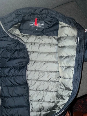 Svart dunjacka från Polo Club - Snygg svart dunjacka från Polo Club med quiltad design och diskret logomärke på ärmen. Jackan har dragkedja framtill och en lätt, puffig känsla som passar perfekt för kyliga dagar. Insidan är ljusgrå och jackan har en modern, stilren look.