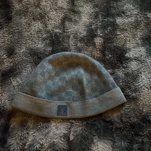 Blå Louis Vuitton mössa - Snygg grå mössa från Louis Vuitton med klassiskt rutmönster och vikt kant. Mössan har en diskret LV-logga framtill och är tillverkad i ett mjukt stickat material som känns lyxigt och bekvämt. Perfekt accessoar för att lyfta din outfit.