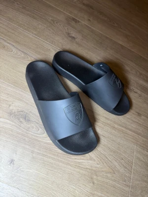 Svarta Lamborghini tofflor - Säljer ett par svarta slides från Lamborghini med klassisk logga på ovandelen. Tofflorna har en enkel och stilren design med bred rem och räfflad sula för extra komfort. Perfekta för chill dagar hemma eller till stranden.