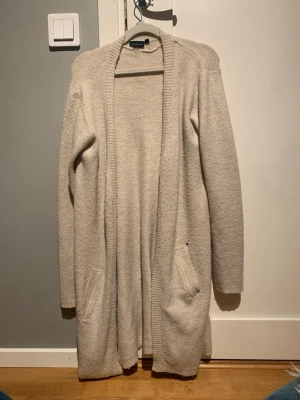 Beige lång kofta  - Mysig lång kofta i beige. Koftan har öppen front, två fickor framtill och ribbade kanter. Perfekt för lager på lager och chill dagar. Materialet är mjukt och lite fluffigt, vilket ger en avslappnad vibe.Har ett hål vid fickan (bild finns)! Därav priset! Super gosigt material!