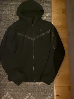 Svart Nike TECH  - Säljer en svart hoodie från Nike med hel dragkedja framtill och huva. Tröjan har en diskret Nike-logga på bröstet och ett stilrent, svart band över bröstet. Praktiska fickor på sidorna och mjukt material som passar perfekt till en avslappnad stil. Kan sänka pris max -100