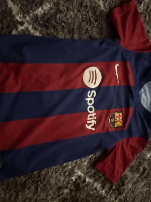 Barcelona matchtröja Nike röd/blå - Säljer en FC Barcelona matchtröja från Nike i klassiska rödblåa ränder. Tröjan har Spotify-logga på bröstet, klubbmärke och Nike swoosh. Tillverkad i lätt och ventilerande material, perfekt för fotboll eller träning. Kortärmad och med UNHCR-logga på ryggen. storlek 137 147