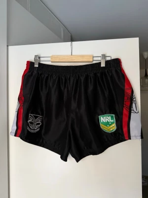 Svarta sportshorts från NRL Warriors - Säljer ett par svarta sportshorts från NRL Warriors med broderade loggor framtill och röda samt vita detaljer längs sidorna. Shortsen har elastisk midja och är perfekta för träning eller sportiga aktiviteter.