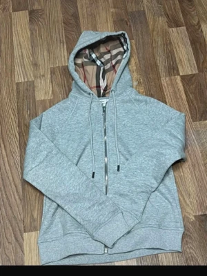 Grå zip hoodie med Burberry-mönster - Köp av mig så fort som möjligt.Snygg grå zip hoodie från Burberry med klassiskt rutigt foder i huvan. Hoodien har dragsko i huvan, långa ärmar och ribbade muddar. Tillverkad i mjukt bomullsmaterial för en skön känsla. Perfekt för dig som gillar stilrena detaljer och ikoniska mönster.