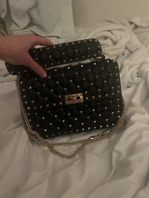 Svart Valentino liknande quiltad handväska med nitar  - Säljer en svart Valentino Liknande quiltad handväska med guldfärgade nitar och kedjerem. Väskan har ett elegant rutmönster, vridlås i guld och en bred topphandtag. Perfekt för dig som vill ha en edgy men stilren accessoar.