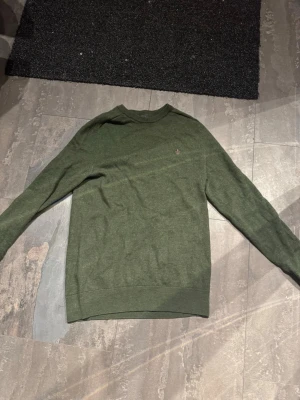 Morris stickad tröja  - Mörkgrön stickad tröja från Morris i 100% merinoull. Tröjan har rund halsringning, långa ärmar och en liten broderad logga på bröstet. Perfekt för lager-på-lager och enkel att matcha med jeans eller chinos.