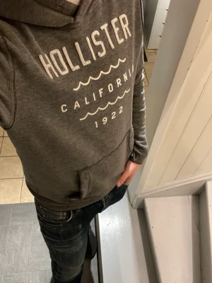 Grå hoodie från Hollister - Grå Hollister hoodie med vitt Hollister California tryck på bröstet. Perfekt för en avslappnad och cool stil. Tröjan har inga defekter förutom att den urtvättad, men det tillför bara en skön vintage känsla på tröjan. Hör av dig vid minsta fundering! 😁