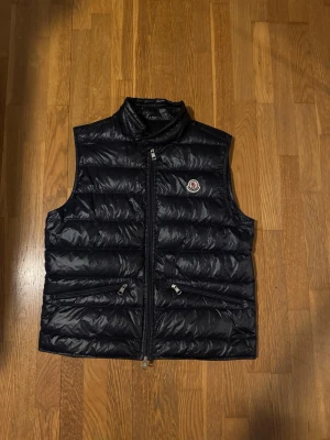 Svart dunväst från Moncler - Snygg svart dunväst från Moncler med hög krage och två dragkedjeförsedda fickor framtill. Västen har Moncler-logga på bröstet och quiltad design som ger en sportig och trendig look. Perfekt för lager-på-lager och streetstyle. Pris kan absolut diskuteras och är öppen för de flesta rimliga priser.