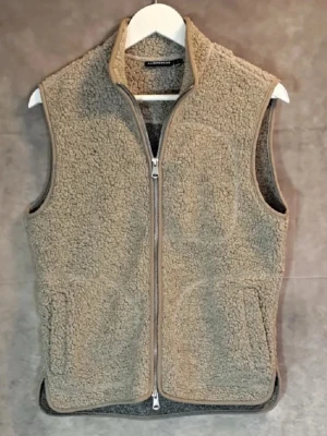 Beige fleeceväst från J.Lindeberg Teddyväst - Mysig beige fleeceväst från J.Lindeberg med hel dragkedja framtill och två stora fickor. Västen har en hög krage och är tillverkad i ett mjukt teddy-material som håller dig varm. Perfekt för lager-på-lager-stil under kyliga dagar.