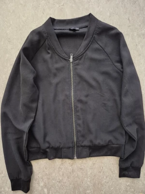 Bomberjacka - En stilren svart bomberjacka med hel dragkedja framtill och ribbade muddar vid ärmslut och midja. Jackan har en klassisk bombermodell med rundad hals och långärmad design. Perfekt för en clean och enkel look. Har en fläck.